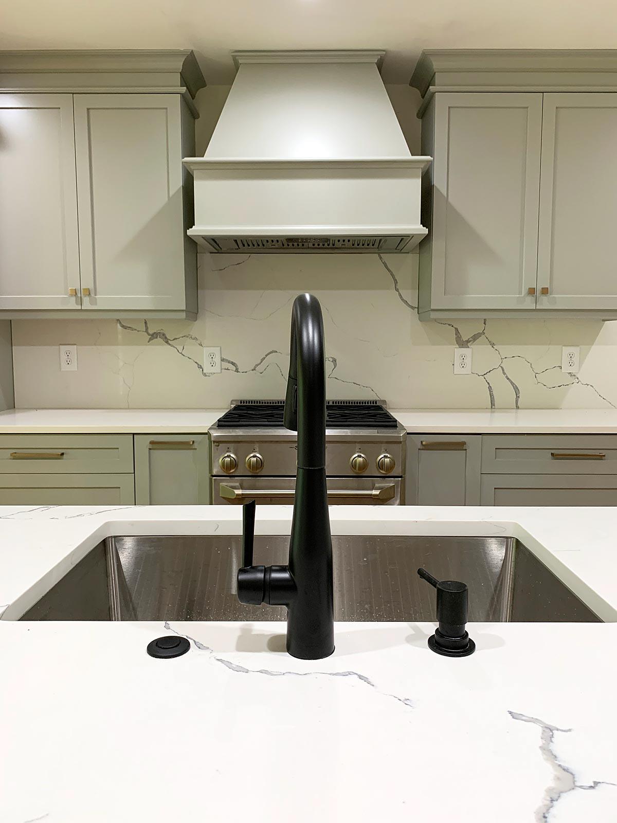 View from kitchen island past sink and looking onto tan cabinetry and gas range with hood fan.