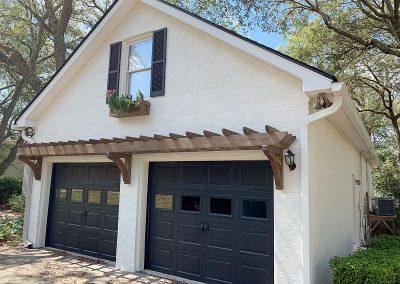 Attached garage, painted white brick with black shutters and garage doors and trellis overhang.