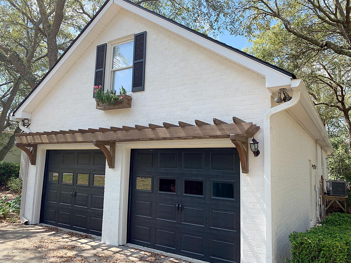 Attached garage, painted white brick with black shutters and garage doors and trellis overhang.