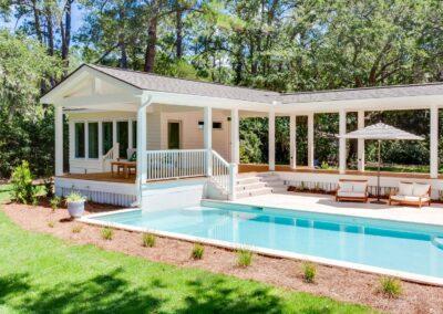 View of pool house from corner of the yard with blue pool and tiled patio in front.