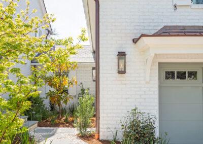 Garage with mother-in-law suite above, painted white brick and copper roof and gutter, paver and pea gravel walkway leading to main house.