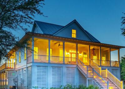 Night time view of white elevated home with multiple roof pitches, deep front porch and small view of the river behind home.