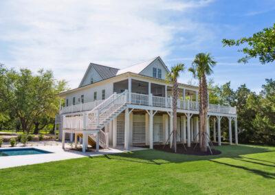 View of back right corner of stately custom home, elevated, painted white and with a pool and landscaping.