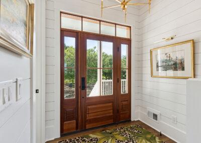 View from inside foyer, looking out of cherry wood front entry door with side lights and multiple pane transom with shiplap, gold accented artwork and light fixture.