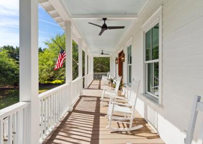Extended view of front porch from one end to the other, several rocking charis and American flag blowing against a backdrop of mature green trees.