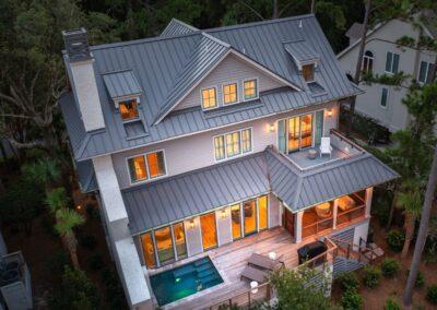 Aerial exterior shot of Kiawah Island custom home from the back showing 3 raised living levels a large deck with pool, screened in porch and an open deck above that sits off of a bedroom. Home is lit and it is around dusk.