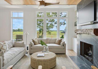 Comfortable living room with high contrast stone fireplace, driftwood mantle, warm wood paneling along the ceiling and linen sofas. Overlooking windows peering out over the marsh.