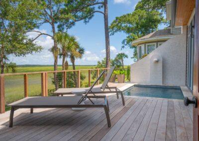 beautiful marsh view from an open air deck with warm wood accents, built-in pool and a modern cable railing overlooking the marsh with pine trees, palmettos and oaks on the property. Architectural detail on the house in the background give interest to the right side of image.