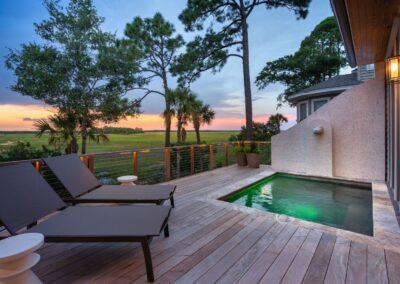 Dusk in the background and image of the emerald green pool on the back deck of a Kiawah Island custom home with two comfy chaise lounge chaires just outside of the built-in deck pool.